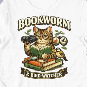 Puede incluir: Camiseta blanca con un gato de dibujos animados con gafas y prismáticos, leyendo un libro. La camiseta tiene el texto "BOOKWORM & BIRD-WATCHER". Un pájaro está posado en una pila de libros.