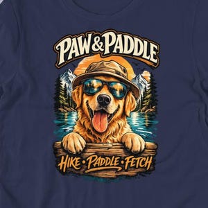 Może przedstawiać: Granatowa koszulka z nadrukiem złotego retrievera w okularach przeciwsłonecznych i kapeluszu, z napisem "PAW & PADDLE" powyżej i "HIKE • PADDLE • FETCH" poniżej. Pies znajduje się w scenerii jeziora.