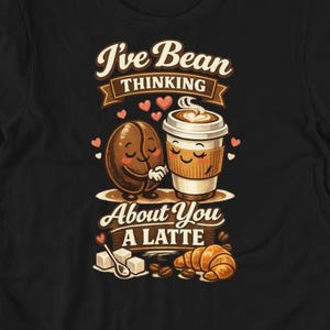 May include: Black t-shirt with a graphic featuring a coffee bean and a coffee cup, both with smiling faces, and the text "I've Bean Thinking About You A Latte". The design includes hearts, sugar cubes, and a croissant.