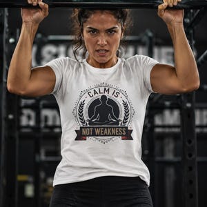 May include: A white short-sleeved t-shirt featuring a black graphic print of a person in a seated meditation pose. The design includes the text "CALM IS NOT WEAKNESS" enclosed within a circular emblem with decorative laurel leaf accents.