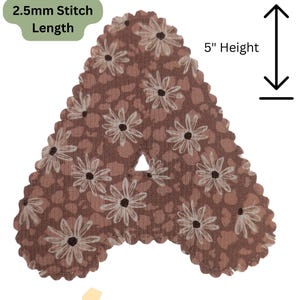 Può includere: Una lettera "A" in tessuto con un motivo floreale marrone e bianco. La lettera è alta 12,7 cm e ha una lunghezza del punto di 2,5 mm. I bordi sono smerlati.