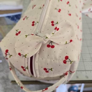 Può includere: Una borsa cilindrica in tessuto con base color crema e un motivo a piccole ciliegie rosse e minuscoli puntini neri. La borsa presenta una chiusura a zip bianca, un dettaglio a fiocco in tessuto sulla parte superiore e un manico abbinato.