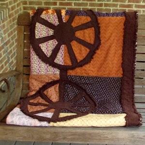 Könnte beinhalten: Eine braune und orangefarbene Patchwork-Decke mit einem Design aus drei braunen Wagenrädern. Die Decke liegt auf einer Holzbank.