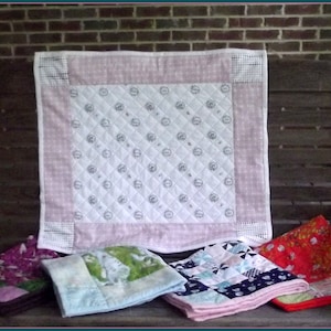 May include: Five different baby quilts, all handmade and featuring various patterns and colors. The quilts are displayed on a wooden bench with a brick wall in the background.