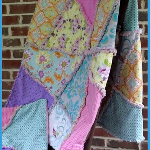 Può includere: Una trapunta patchwork con una varietà di motivi floreali e geometrici in tonalità di rosa, blu, viola e giallo. La trapunta è realizzata in tessuto morbido e testurizzato e ha un bordo arricciato.