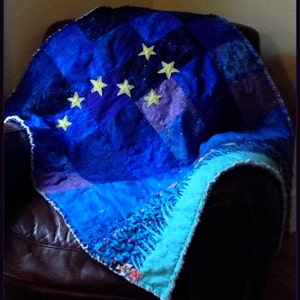 Può includere: Una trapunta patchwork blu e viola con un bordo bianco e applicazioni di stelle gialle. La trapunta è drappeggiata su una poltrona in pelle marrone.