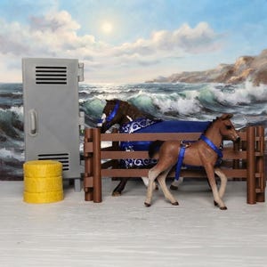 May include: A brown horse figurine wearing a blue blanket and a smaller brown foal figurine with a blue harness standing behind a brown fence. Next to them is a gray locker and a stack of three yellow cylindrical items.