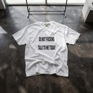 May include: A white short-sleeve t-shirt featuring the black text "DO NOT FUCKING TALK TO ME TODAY" printed in a bold, sans-serif font across the center of the chest. The garment has a crew neckline and a standard fit with visible fabric folds.
