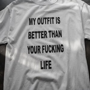 May include: A white short-sleeved t-shirt featuring bold black text printed on the front that reads, MY OUTFIT IS BETTER THAN YOUR FUCKING LIFE. The garment has a standard crew neckline and is constructed from a plain, solid-colored fabric.