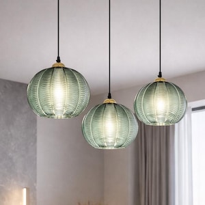 May include: Three pendant lights with ribbed, seafoam green glass shades hang above a modern living room. The shades have a round shape and are suspended from black cords with gold-colored accents. The room features a neutral color palette with a large sectional sofa.