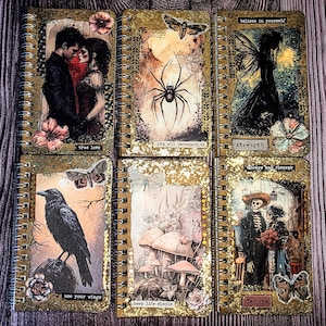 May include: Six spiral-bound notebooks with gold glitter covers and varied collage art. Designs include a couple, a spider, a fairy, a raven, mushrooms, and skeletons. Text includes "true love," "it's all perspective," "strength," "use your wings," "keep life simple," and "always and forever."