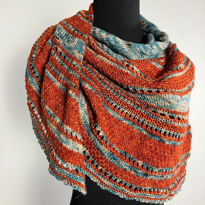 May include: A knitted shawl with a rust and blue color scheme. The shawl is made with a delicate stitch pattern and has a fringed edge.