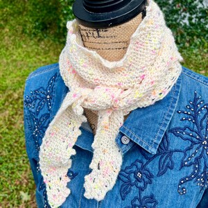 May include: A cream-colored knit scarf featuring a speckled pattern of pink, yellow, and orange flecks. The scarf has a textured, open-stitch design with pointed, lace-like edges and is tied in a simple knot around the neck of a display form.