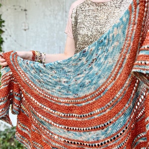 Puede incluir: Un chal de punto de color azul verdoso y naranja con un patrón circular. El chal está hecho con un hilo suave y tiene un delicado diseño de encaje.