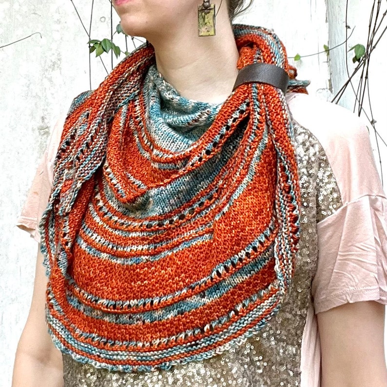 May include: A knitted shawl in shades of orange, blue, and white with a decorative crocheted border. The shawl is worn around the neck of a person.