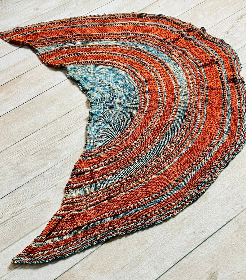May include: A hand-knitted shawl in shades of blue and orange with a circular pattern. The shawl is made with a soft yarn and has a delicate texture.