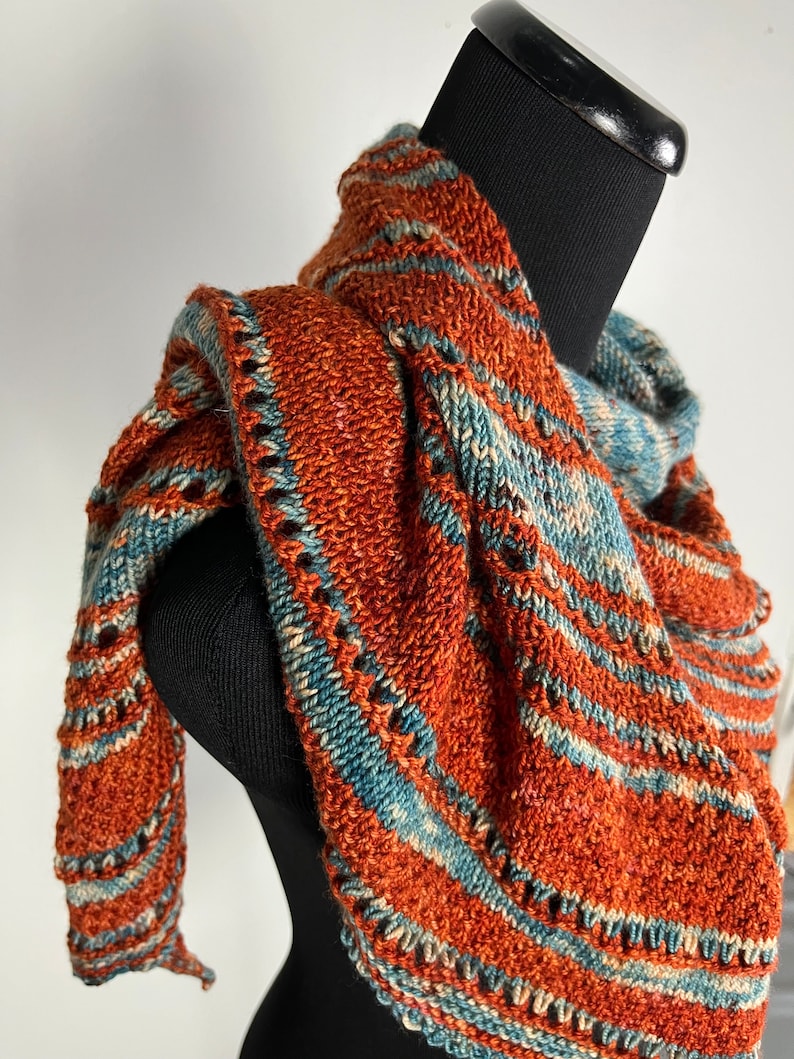 May include: A knitted shawl with a triangular shape, featuring a rust orange and teal blue color scheme. The shawl is made with a textured stitch and has a fringe border.