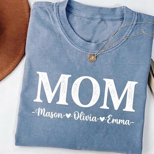 Könnte beinhalten: Ein blaues T-Shirt mit dem Wort "MOM" in großen weißen Buchstaben aufgedruckt. Unter dem Wort "MOM" stehen die Namen "Mason", "Olivia" und "Emma", getrennt durch Herzen. Eine silberne Halskette mit rundem Anhänger liegt über dem Shirt.
