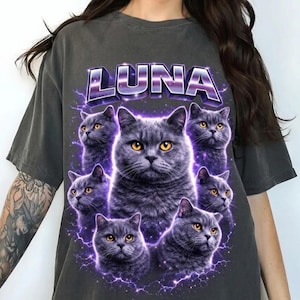 Peut inclure: T-shirt gris foncé avec un motif de plusieurs chats gris aux yeux jaunes, entourés d'éclairs violets. Le mot "LUNA" est imprimé au-dessus des chats en violet et argent.