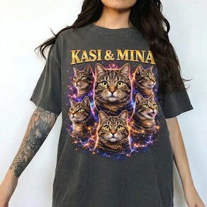 Può includere: T-shirt grigio scuro con una grafica di diversi gatti tabby con occhi gialli, circondati da un design cosmico. Il testo "KASI & MINA" è stampato in lettere dorate sopra i gatti.