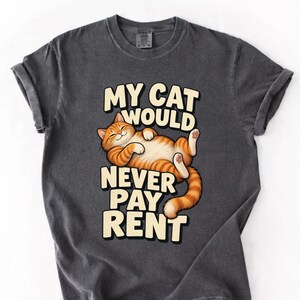 May include: A charcoal gray t-shirt featuring a graphic of a relaxed orange tabby cat lying on its back. The text "MY CAT WOULD NEVER PAY RENT" is printed in large, cream-colored block letters above and below the cat illustration.