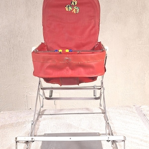 May include: A red high chair with a metal frame and a fabric seat featuring an embroidered design of alphabet blocks. The seat includes a horizontal bar with small, colorful beads. The structure is supported by a silver-colored metal base.