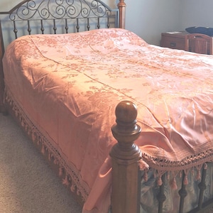 May include: A bed with a wrought iron headboard and footboard. The bed is covered with a peach-colored floral patterned bedspread and a matching bed skirt with tassels. The bed frame is made of wood.