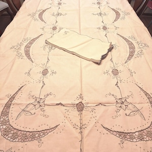 May include: A rectangular cream-colored tablecloth featuring intricate brown embroidery of floral motifs and crescent-shaped cutwork patterns. A stack of matching cream-colored napkins with embroidered edges rests in the center of the tablecloth.