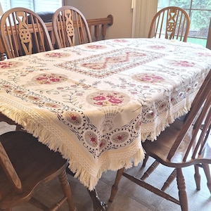 May include: Cream-colored oval tablecloth with a floral pattern in pink, red, and gray. The tablecloth has a fringe trim and is set on a wooden table surrounded by wooden chairs. The floral design includes bouquets and fan-shaped elements.