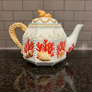 May include: A white ceramic teapot with a seashell and coral design. The teapot features a gold handle and accents, with red coral and seashell details. The lid is topped with a seashell. The teapot is approximately 10 inches tall.