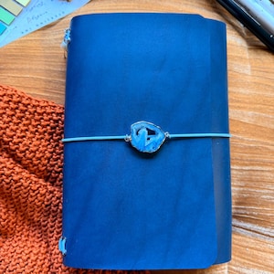 May include: A blue leather journal with a decorative blue stone clasp and a light blue elastic closure. The journal is made of Buttero Italian leather. The word "Blue" is printed at the bottom of the image.