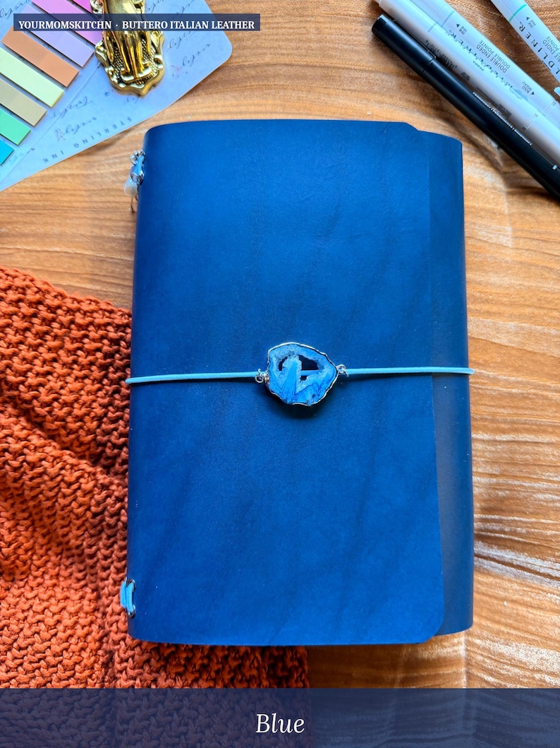 May include: A blue leather journal with a decorative blue stone clasp and a light blue elastic closure. The journal is made of Buttero Italian leather. The word "Blue" is printed at the bottom of the image.