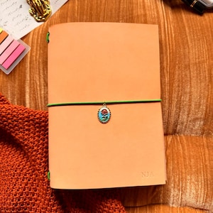May include: A natural-colored leather journal with a green elastic closure and a decorative charm. The journal is personalized with the initials "NJA". The text "YOURMOMSKITCHN - BUTTERO ITALIAN LEATHER" is at the top. The word "Natural" is at the bottom.