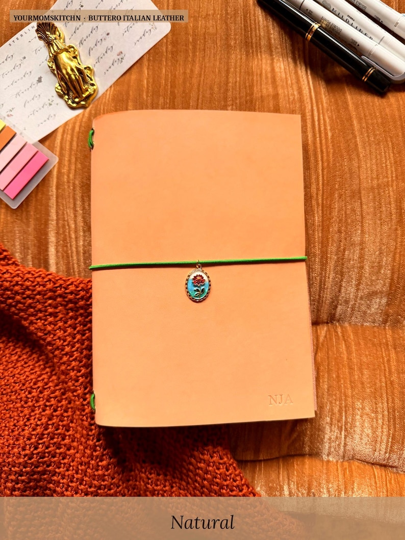 May include: A natural-colored leather journal with a green elastic closure and a decorative charm. The journal is personalized with the initials "NJA". The text "YOURMOMSKITCHN - BUTTERO ITALIAN LEATHER" is at the top. The word "Natural" is at the bottom.