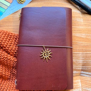 May include: A mahogany-colored leather journal with a sun charm and elastic closure. The journal has the initials "M.C.P." embossed on the front. The text "YOURMOMSKITCHN - BUTTERO ITALIAN LEATHER" is at the top.