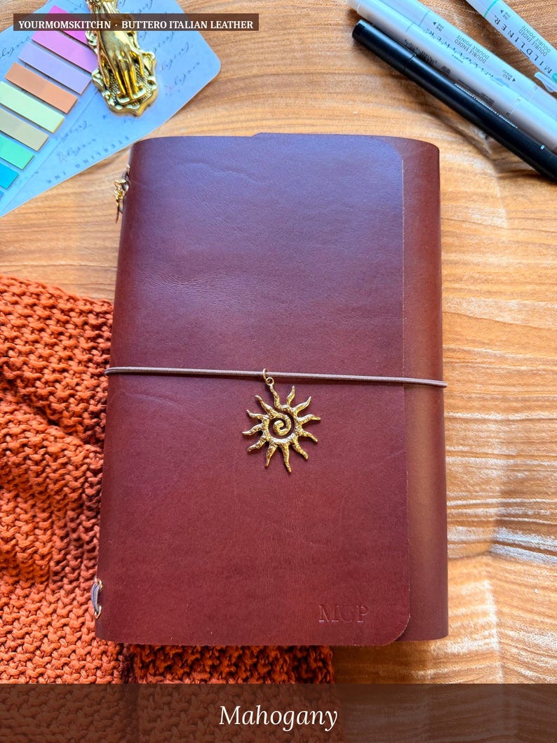 May include: A mahogany-colored leather journal with a sun charm and elastic closure. The journal has the initials "M.C.P." embossed on the front. The text "YOURMOMSKITCHN - BUTTERO ITALIAN LEATHER" is at the top.