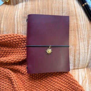 May include: A deep burgundy leather journal featuring a smooth, matte finish. A thin black elastic band wraps horizontally around the center, secured with a small, metallic gold-toned sun charm that hangs from the band at the front cover.