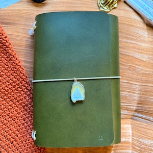 May include: Olive green leather journal with a decorative charm and elastic closure. The journal is made of Italian leather and features the text "YOURMOMSKITCHN - BUTTERO ITALIAN LEATHER".