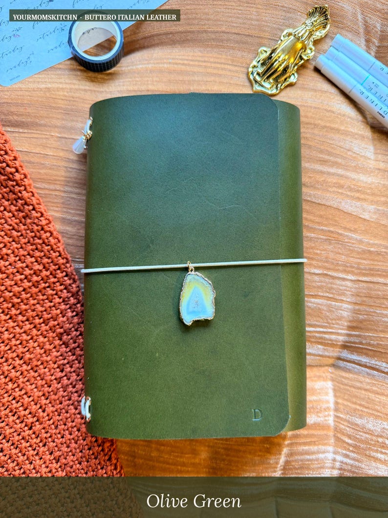 May include: Olive green leather journal with a decorative charm and elastic closure. The journal is made of Italian leather and features the text "YOURMOMSKITCHN - BUTTERO ITALIAN LEATHER".