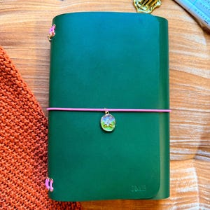 May include: Emerald green leather journal with a pink elastic closure and a decorative charm. The journal has the initials "JMB" embossed on the cover. The text "YOURMOMSKITCHN - BUTTERO ITALIAN LEATHER" is at the top. The text "Emerald Green" is at the bottom.