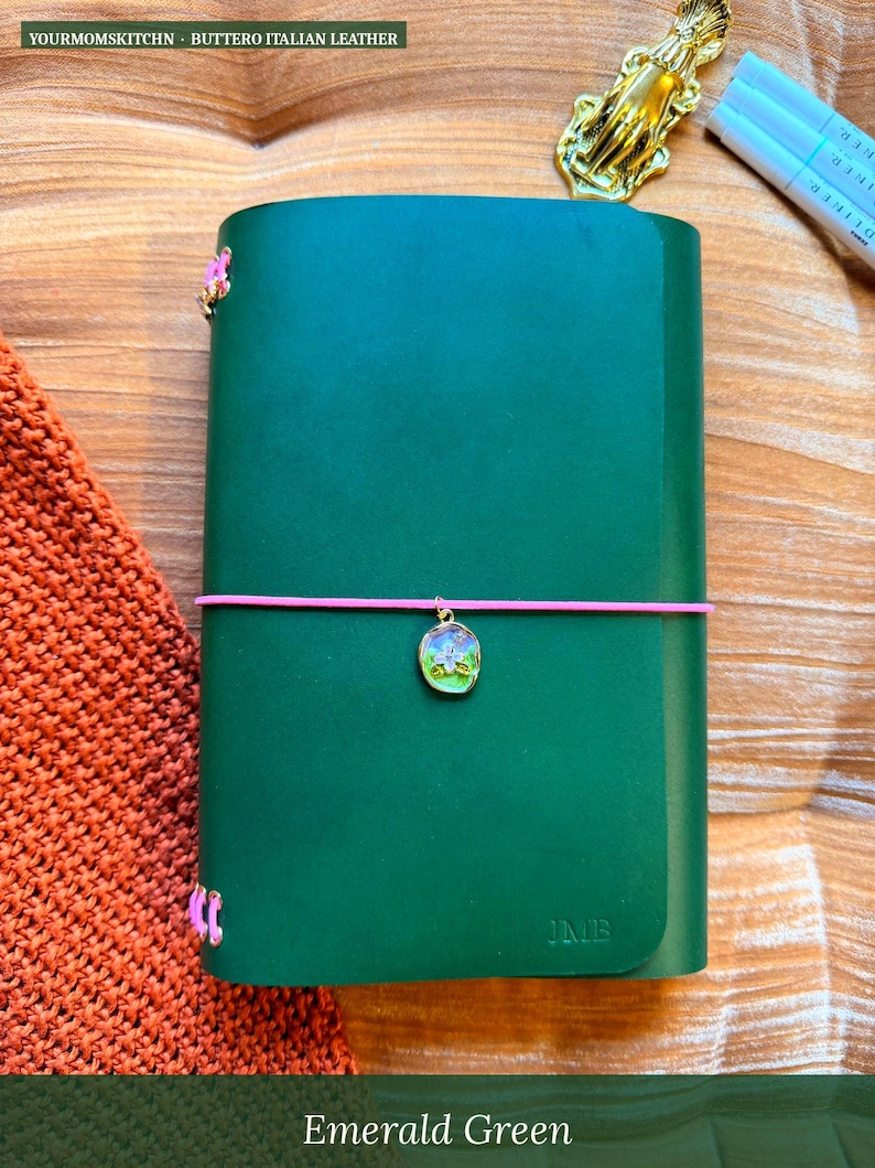 May include: Emerald green leather journal with a pink elastic closure and a decorative charm. The journal has the initials "JMB" embossed on the cover. The text "YOURMOMSKITCHN - BUTTERO ITALIAN LEATHER" is at the top. The text "Emerald Green" is at the bottom.