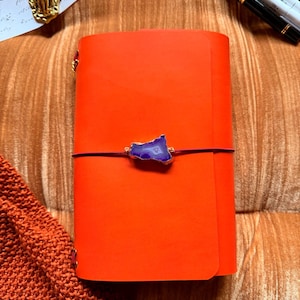 May include: An orange leather journal with a purple stone accent and a dark purple elastic closure. The journal is closed and has gold-colored corner protectors. The text "YOURMOMSKITCHN - BUTTERO ITALIAN LEATHER" is at the top.