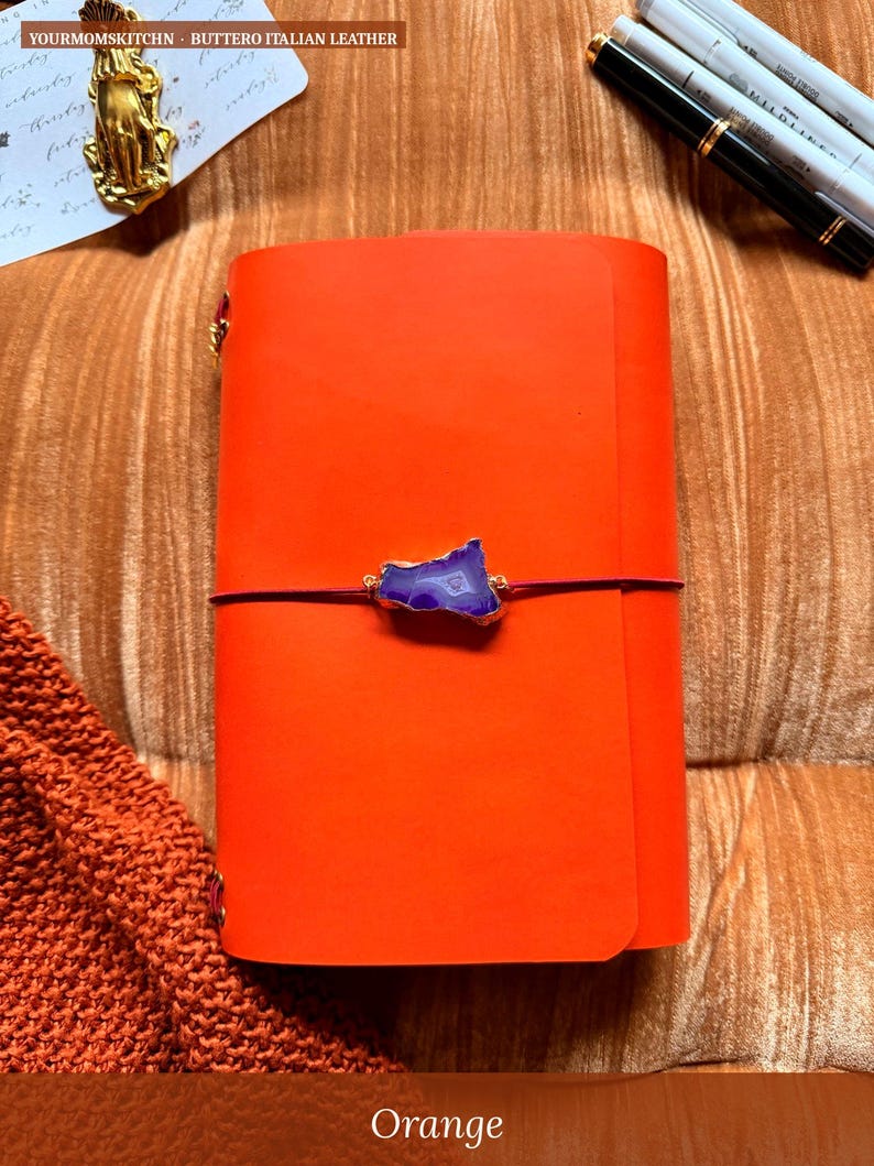 May include: An orange leather journal with a purple stone accent and a dark purple elastic closure. The journal is closed and has gold-colored corner protectors. The text "YOURMOMSKITCHN - BUTTERO ITALIAN LEATHER" is at the top.