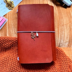 May include: A chestnut-colored leather journal with a decorative charm and elastic closure. The journal is embossed with the initials "SS" and the text "YOURMOMSKITCHN - BUTTERO ITALIAN LEATHER" is visible at the top. The word "Chestnut" is at the bottom.