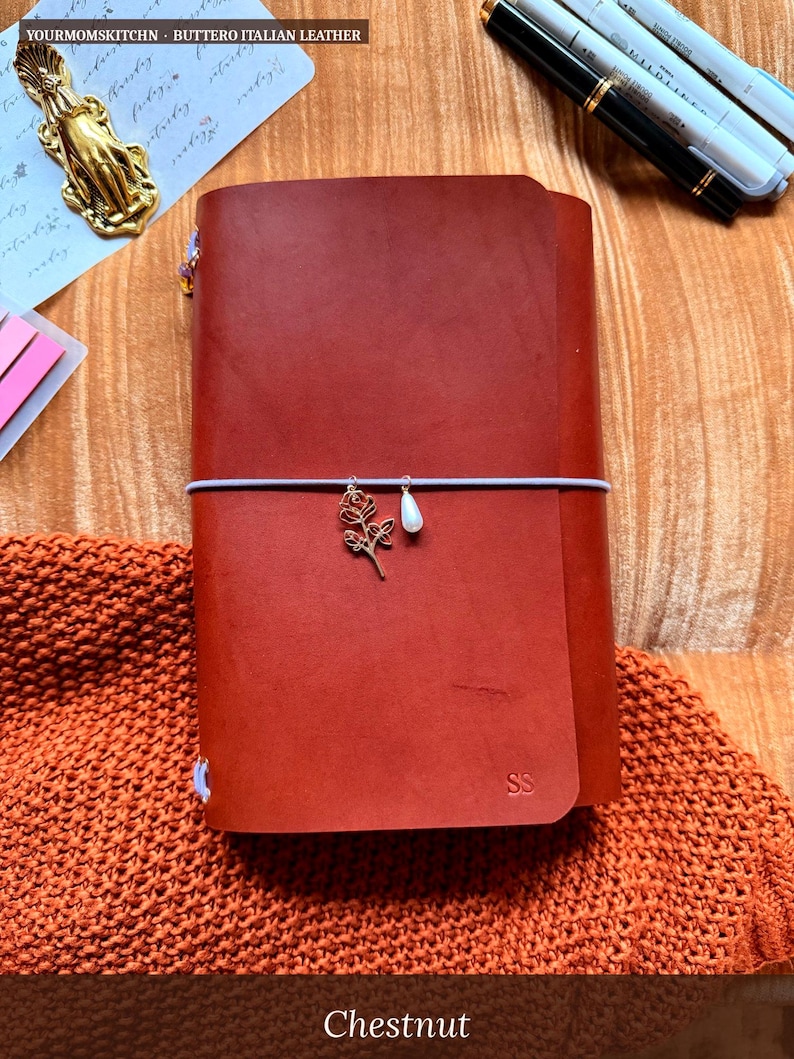 May include: A chestnut-colored leather journal with a decorative charm and elastic closure. The journal is embossed with the initials "SS" and the text "YOURMOMSKITCHN - BUTTERO ITALIAN LEATHER" is visible at the top. The word "Chestnut" is at the bottom.