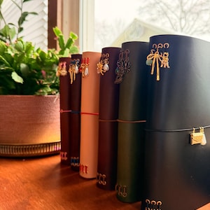 May include: A row of leather journals in various colors, each adorned with decorative charms. The journals are secured with elastic closures and feature gold-toned metal accents. A potted plant sits in the background.