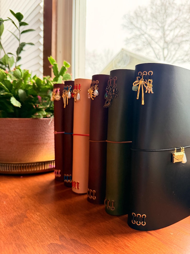 May include: A row of leather journals in various colors, each adorned with decorative charms. The journals are secured with elastic closures and feature gold-toned metal accents. A potted plant sits in the background.