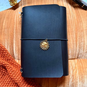May include: Black leather journal with a gold sun charm and elastic closure. The journal is made of Buttero Italian leather. The text "Black" is visible at the bottom of the image.