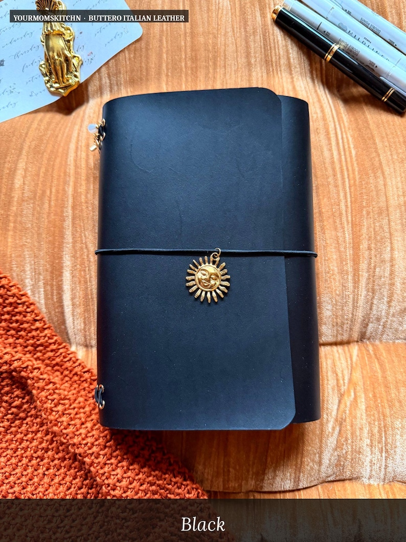 May include: Black leather journal with a gold sun charm and elastic closure. The journal is made of Buttero Italian leather. The text "Black" is visible at the bottom of the image.