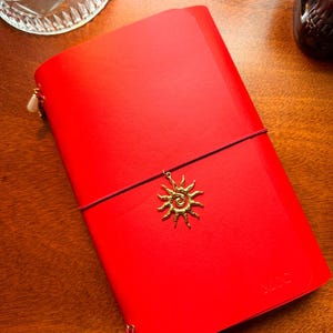 May include: A bright red leather journal with a sun charm and elastic closure. The journal has a gold-colored charm and the word "Red" is at the bottom of the image.